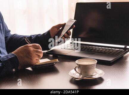 Casual businessman with smartphone and laptop working from home writing in his notebook with a cup of coffee on the table on a sunny day Stock Photo