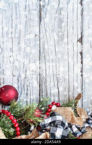 Snowy Christmas background with holiday trimmings of pine tree branches, ornaments, black and white buffalo check ribbon, burlap and red bead garland. Stock Photo