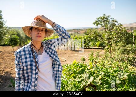 Portrait of young farmer woman with straw hat and blue checkered shirt in the vegetable garden resting and taking air Stock Photo