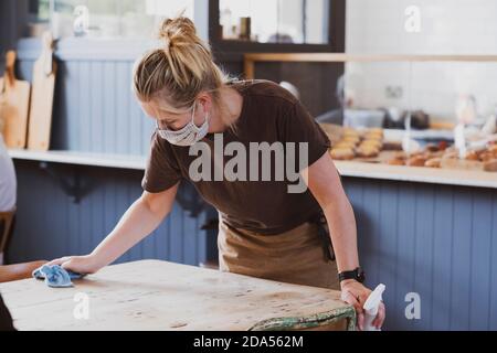 Blond waitress wearing face mask working in a cafe, wiping table. Stock Photo