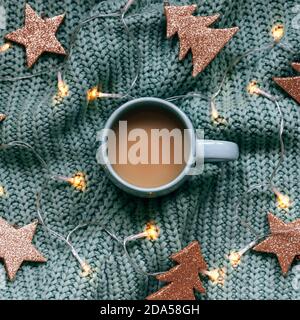 A cup of coffee on knitted sweater with garland lights and Christmas decor, top view Stock Photo
