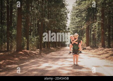 Adorable blonde boy carrying his little brother on his back and posing in the middle of a footpath Stock Photo