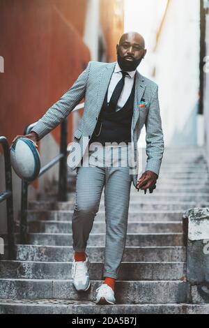 A serious bald bearded fancy black businessman in a fashionable plaid suit with a necktie and orange socks is descending outdoor stairs in some Europe Stock Photo