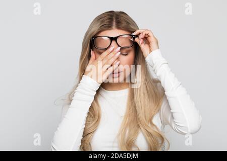 Closeup portrait of sleepy young business woman in glasses in white top rubbing her eyes, feels tired after working on a laptop, front view/Overwork, Stock Photo