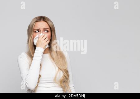 Studio portrait of cute unhealthy blonde female in white top with napkin blowing nose, looking aside. Sick desperate female has flu. Rhinitis, cold, s Stock Photo