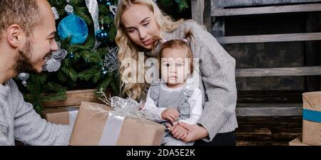 Happy parents give a Christmas gift to a child in front of Christmas tree. Baby girl looks at the camera. Holidays, presents, christmas, x-mas, birthd Stock Photo