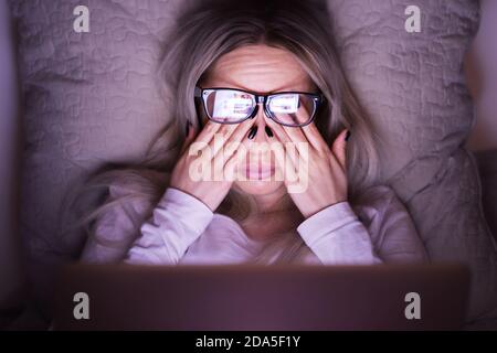 Sleepy young caucasian woman in glasses rubbing her eyes, feels tired after working on a laptop, lying on pillow, front view/ Overwork, tired, health Stock Photo