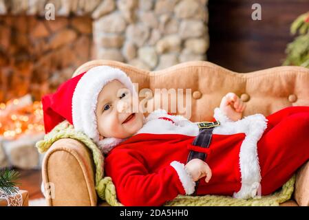 Close-up portrait of a Little boy dressed as red Santa Claus, playing by stone fireplace with bright garlands, putting a gift box under Christmas tree Stock Photo