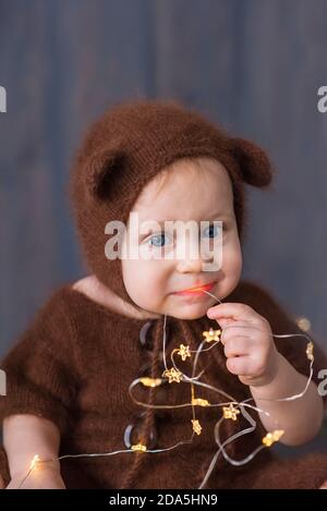 Happy little boy in a brown, fluffy, knitted bear costume, sits on the furon the wooden floor, plays with a bright Christmas garland of lights. Stock Photo