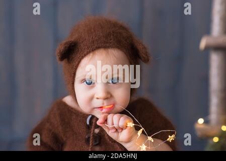 Happy little boy in a brown, fluffy, knitted bear costume, sits on the furon the wooden floor, plays with a bright Christmas garland of lights. Stock Photo