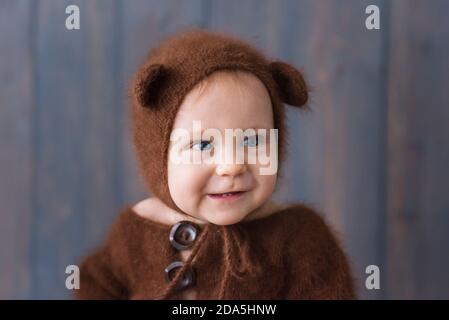 Happy little boy in a brown, fluffy, knitted bear costume, sits on the furon the wooden floor, plays with a bright Christmas garland of lights. Stock Photo