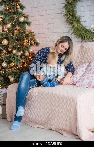 A beautiful young woman sits on a sofa with a boy near Christmas tree against a white brick wall. Mother hugs, kisses, plays with her small son. Happy Stock Photo