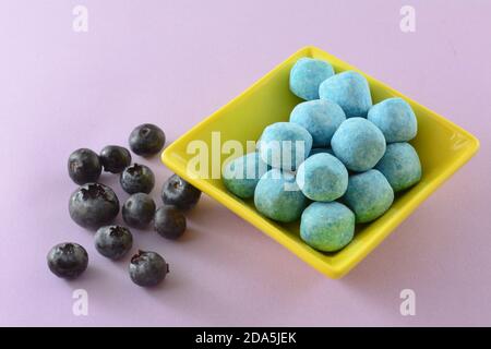 Sour blueberry bonbon candy in yellow candy dish with fresh raw blueberries on lavender background Stock Photo