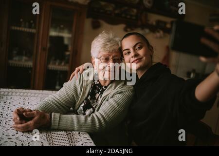 A caucasian grandmother with glasses and her teenage granddaughter take a selfie on the phone while sitting at a table in the house Stock Photo