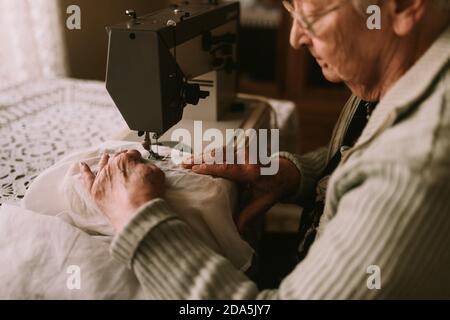 Close up of a senior caucasian woman with glasses sewing a curtain on a sewing machine on a table in the house. Hobbies of senior women Stock Photo