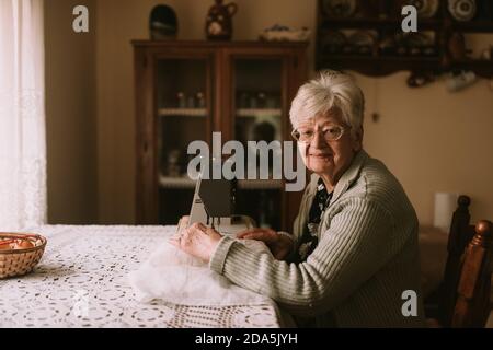 Portrait of a caucasian grandmother with retired glasses who sews a curtain on a sewing machine on the table. Retired old ladies hobby Stock Photo