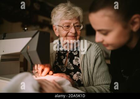 Senior cute smiling grandmother looks at her granddaughter as she teaches her to sew on a sewing machine in the living room Stock Photo