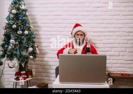 Santa Claus is sitting in the house and pointing to a bottle of champagne while talking on a video call via laptop. Santa Claus online celebrates Stock Photo