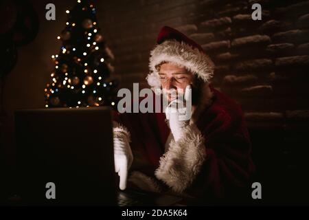 Santa Claus is sitting in front of a laptop in a dark room, while the lights from the Christmas tree can be seen in the background. Christmas and New Stock Photo