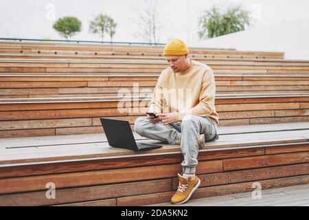 Stylish man hipster in yellow hat freelancer working on laptop in the street city park Stock Photo