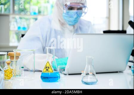 Lab equipment with blue liquid inside stand on the table with male scientist wearing protection suit working with computer laptop for research at labo Stock Photo