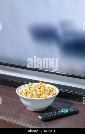 A whtie bowl full of popcorn in front of a led tv with a remote control. Stock Photo