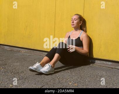 beautiful woman relaxing after outdoors workout. sporty girl getting rest after training. outdoors training, running and sport concept. woman sitting near yellow wall with closed eyes on a stadium. Stock Photo
