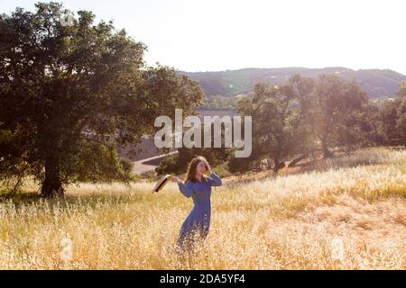 Beautiful smiling young woman standing in a relaxed pose standing in a meadow of gold grasses with Oak trees and farm lands in background Stock Photo