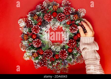 one female hand in knitted sweater adds fir cone to Christmas wreath of cones, berries and balloons on a red background Stock Photo