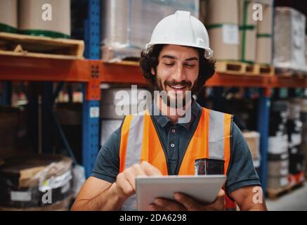 Cheerful young caucasian smiling worker with helmet on head using tablet for work while standing in warehouse against goods Stock Photo