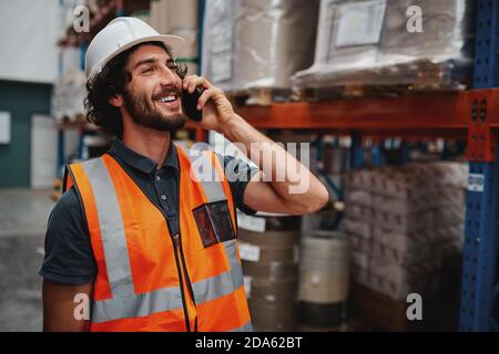Happy bearded warehouse manager in conversation on mobile phone wearing white helmet and safety vest standing looking away Stock Photo