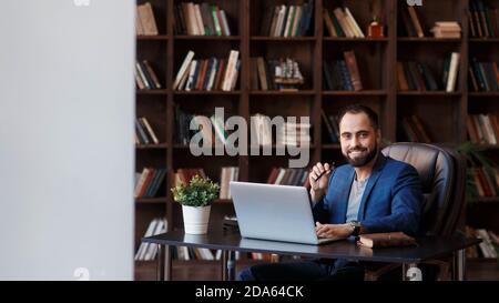 A young bearded businessman in the library office is sitting at a Desk . a man is working on a laptop Stock Photo