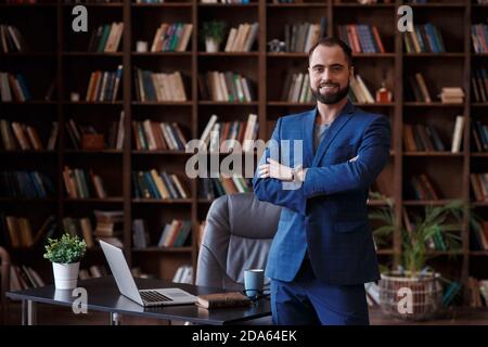 Young bearded businessman in a blue suit in the office library. A man stands at the desktop with a laptop Stock Photo