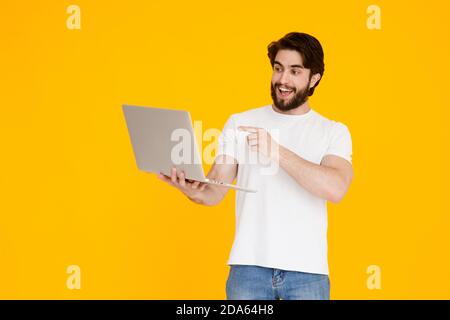 Portrait of a smiling young handsome bearded man holding laptop computer while looking and pointing on screen in isolated studio yellow background Stock Photo