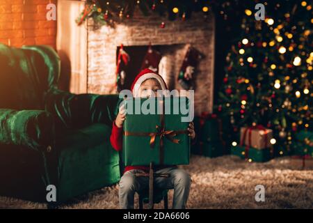 The boy sits on a wooden swing in the shape of a horse. Christmas mood. In the background a decorated Christmas tree and a large fireplace Stock Photo