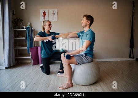 Female nurse helping young man doing stretching exercise in hospital using dumbbells Stock Photo