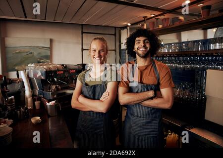 Portrait of young female and male staff wearing apron standing behind counter in cafe with crossed hands looking at camera Stock Photo