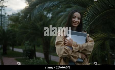 College student woman in coat with laptop and documents in hands, looking at camera on nature background and smiling, education and technology concept Stock Photo