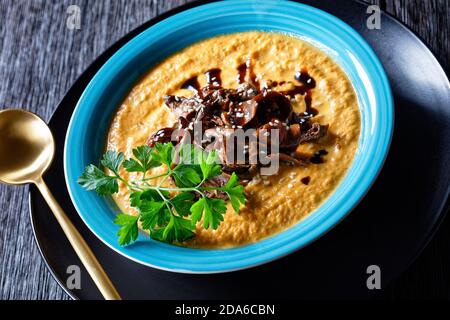 Pumpkin soup of roasted pumpkin puree and wild mushrooms broth, topped with sesame seeds, vegan comfort dish, served on a blue plate with spoon and ga Stock Photo