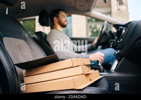 Close-up of bearded delivery man wearing casual clothes driving car delivered hot pizza to customer. Stock Photo