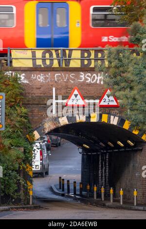 Lower Downs Road, Wimbledon, London UK. 10 November 2020. A Network Rail report shows this Wimbledon railway bridge as one of the “most bashed” in London, coming in at 2nd and struck 11 times in 2019-20. It is also 10th equal most bashed in Britain. Above this narrow arched bridge are four rail tracks, two for commuter trains and two for express trains to and from London Waterloo to the south west of England. The Low Bridge parapet sign is partly obscured by tree foliage on the southern approach. Credit: Malcolm Park/Alamy Live News. Stock Photo