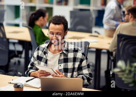 Young happy mixed race man reading notes while working in the modern office, selective focus. Business and startup concept Stock Photo