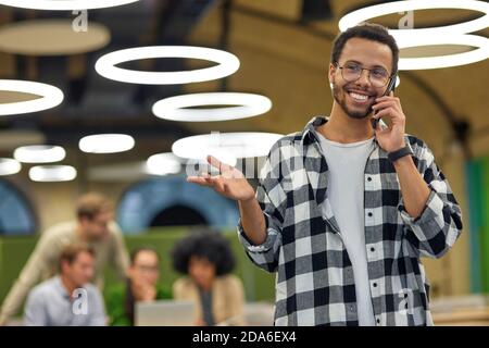 Young happy mixed race man talking by phone and smiling while standing in coworking space with colleagues working on the background. Business people and modern technologies Stock Photo