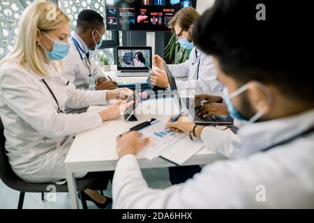 Online video conference, telemedicine, webinar meeting. Team of multiethnic physicians wearing masks, using laptop and tablets, listening the speech Stock Photo