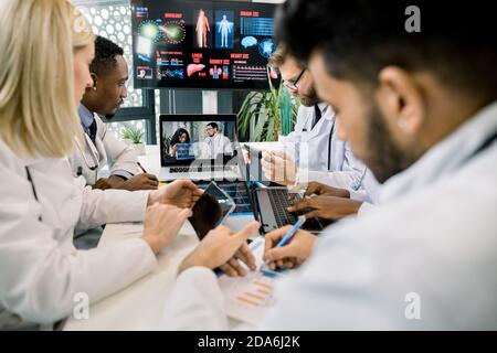 Team of five diverse multiethnic doctors having video conference meeting in hospital, chatting with their two young African and Caucasian colleagues Stock Photo