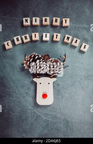 Merry Christmas poster with wooden rain deer,  red nose, pinecones and Christmas lights on a grey background. Christmas mood. Christmas greeting card. Stock Photo
