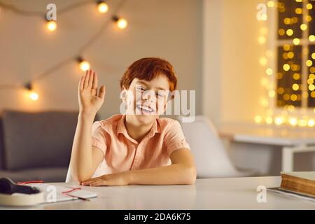 Happy kid waving hand, smiling and looking at camera sitting at desk in cozy room with fairy lights Stock Photo