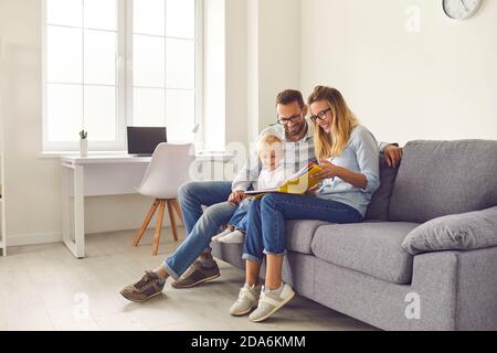 Happy parents and kid sitting on sofa, reading book of stories or looking through family photo album Stock Photo