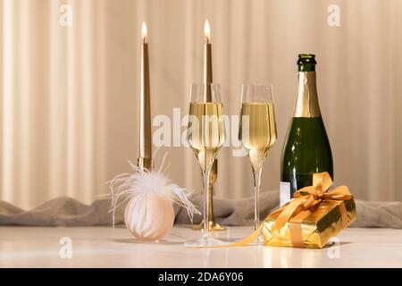 Two champagne glasses surrounded by a Christmas bauble, a wrapped gift box, a champagne bottle and candles on a table. Christmas celebration concept. Stock Photo
