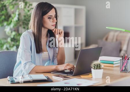 Photo of pensive young lady sit desktop finger chin look laptop calm face wear eyeglasses shirt in home office indoors Stock Photo
